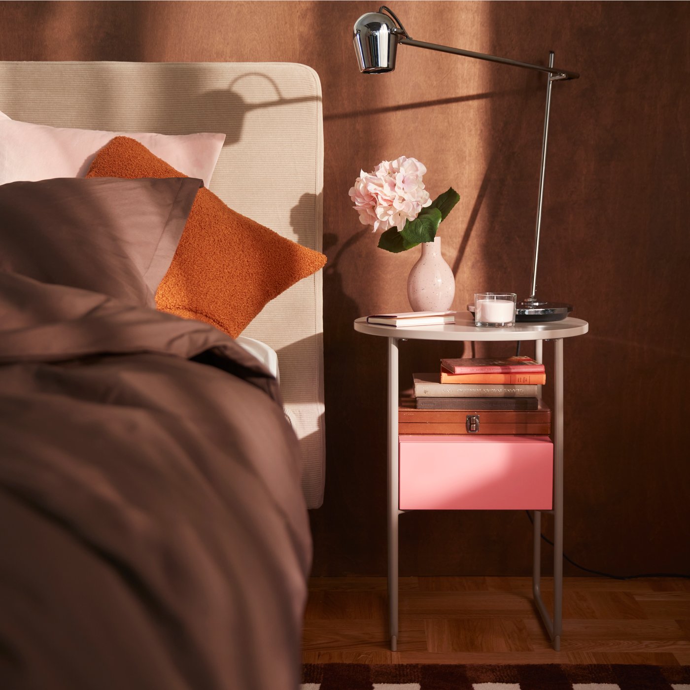Ein Schlafzimmer mit einem beige-rosa BLANKHÄLLAN Nachttisch neben einem Bett. Auf dem Nachttisch stehen Blumen und eine Lampe, vor einer braunen Wand.