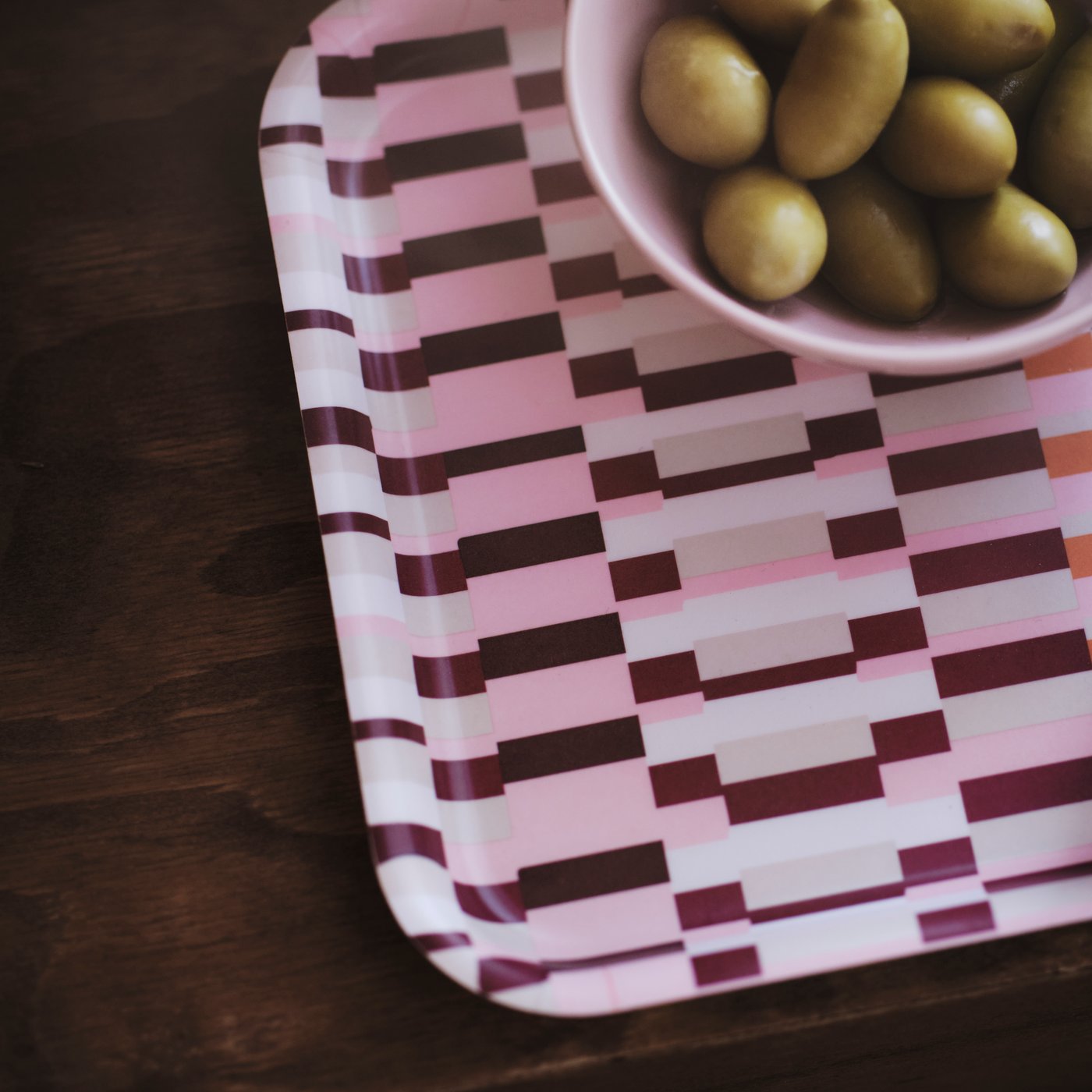 Close-up of the pink patterned HÖSTAGILLE tray with a bowl of green olives on a dark wooden table.
