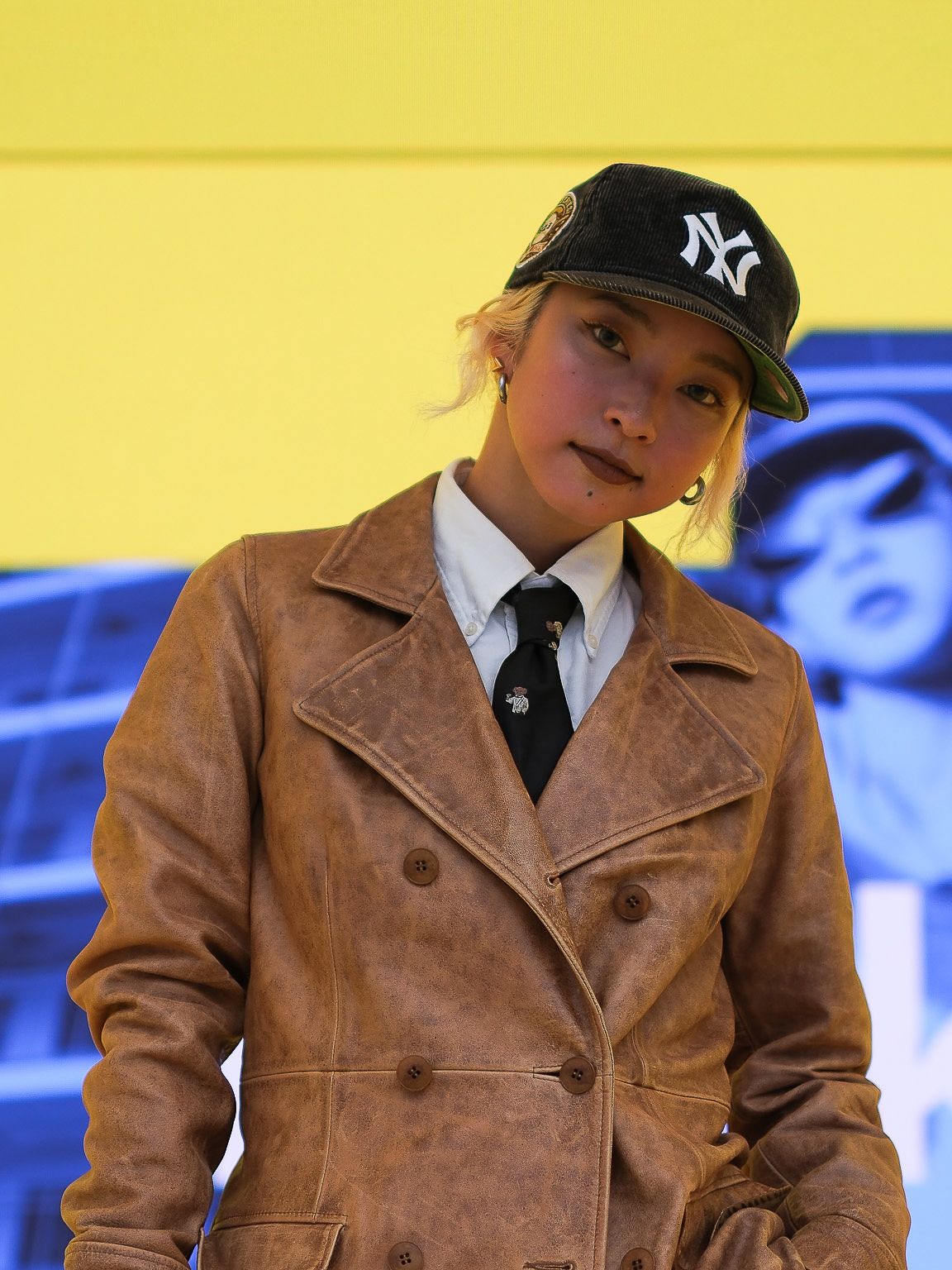 Kyoka staat op een podium in een leren jas, pet, stropdas en wit hemd, met achter haar een foto van haarzelf in blauw op een gele achtergrond afkomstig van de digitale display die tijdens de designweek in Milaan editie 2024 werd gebruikt in het IKEA paviljoen. 