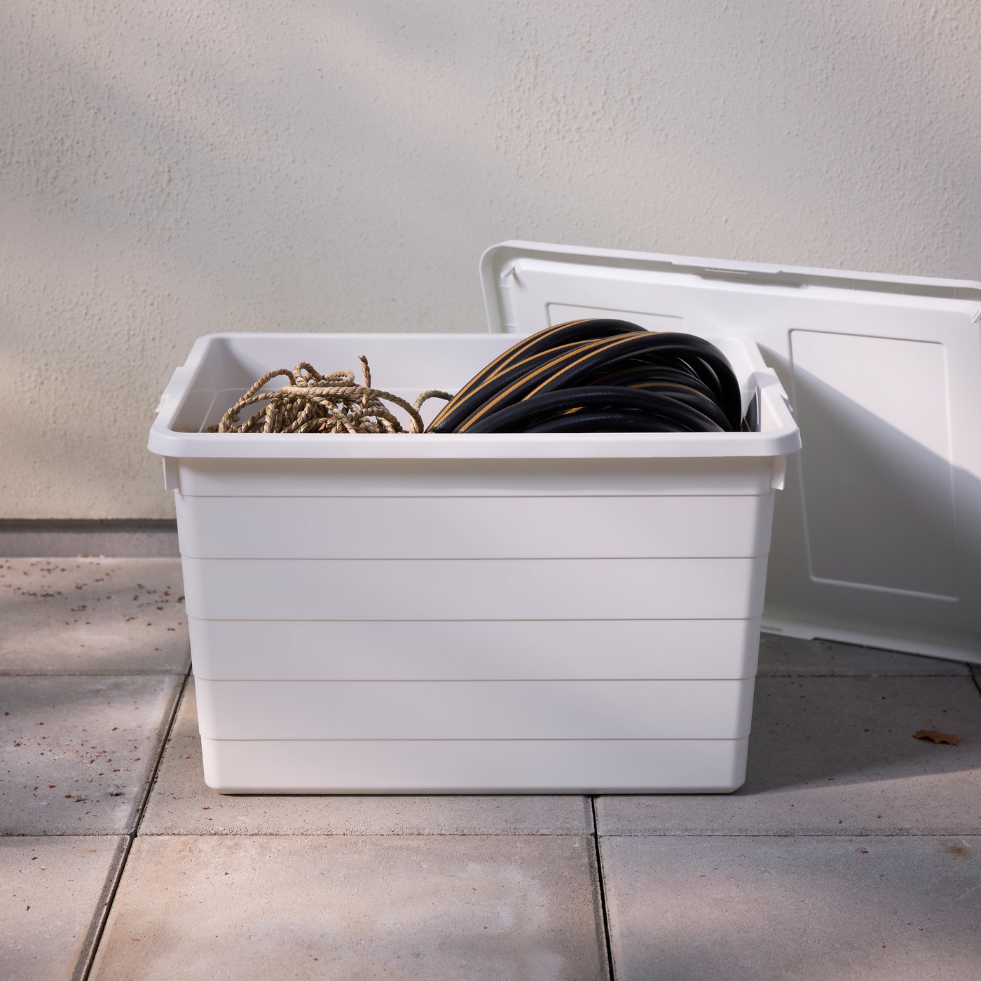 Una caja SOCKERBIT blanca con tapa que contiene un trozo de cuerda y una manguera de jardín en una terraza en un día soleado.