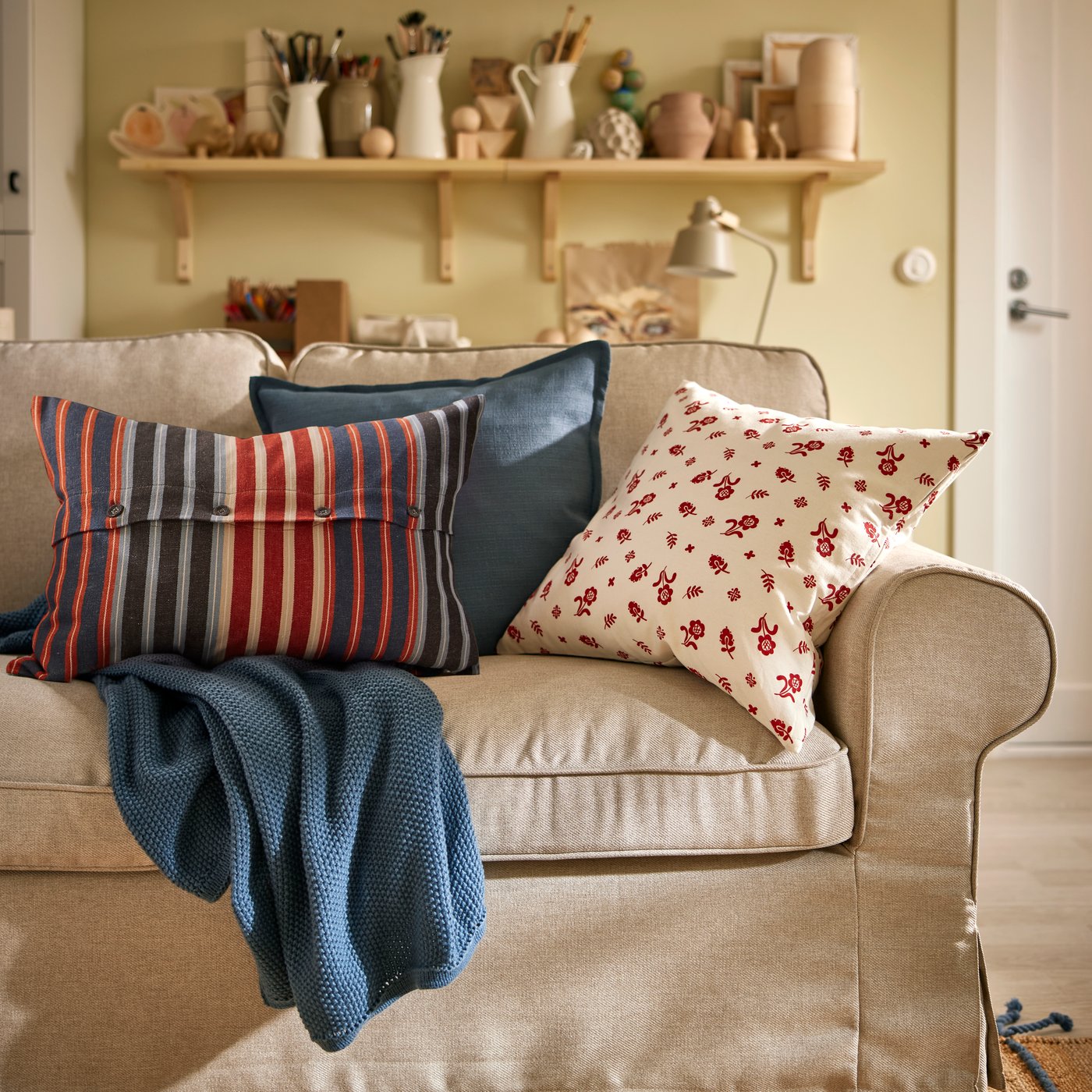 På EKTORP-soffan ligger en blågrå HUMLEMOTT pläd samt några kuddar, däribland en med det blommiga TÅGSÄCKMAL kuddfodralet.