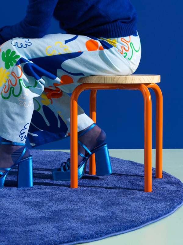 A person in trousers made from KRYPKORNELL pre-cut fabric with a leaf pattern sits on a DOMSTEN stool with orange legs.