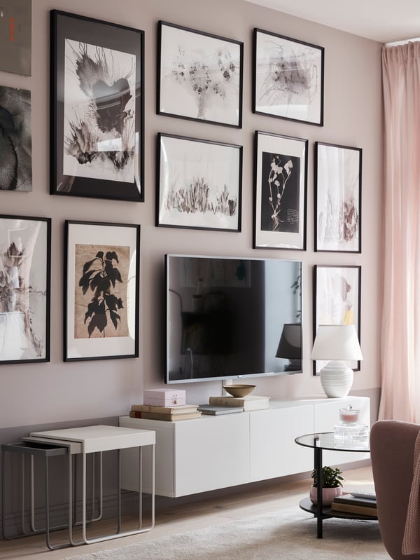 Una pared con muchos cuadros decorativos colgados. Delante de la pared hay unas mesas laterales apilables y al costado un rack de TV con un televisor encima.