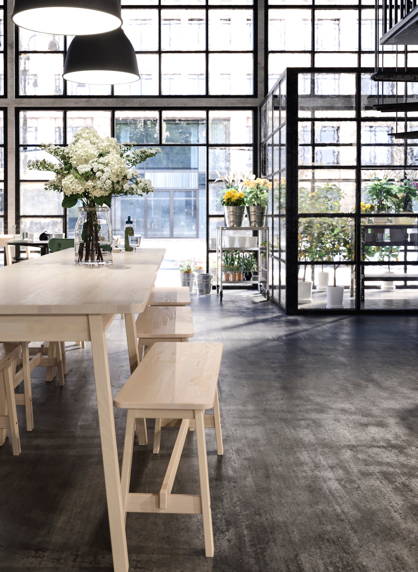 Bright, modern room with large barred windows, a long wooden table with benches and flower arrangements, black pendant lamps above and a glassed-in, secluded area with plants in the background.