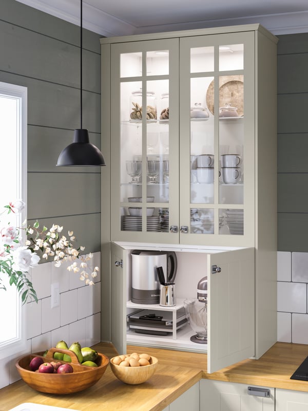 A cabinet on top of a wooden countertop with tableware and appliances. Bowls of fruit and a black pendant lamp.