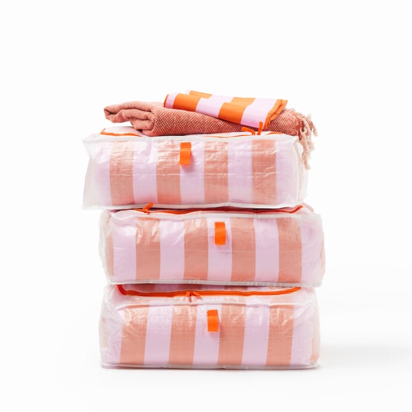A stack of 3 white PÄRKLA storage cases with orange zippers and tabs. The cases are filled with orange striped linens.