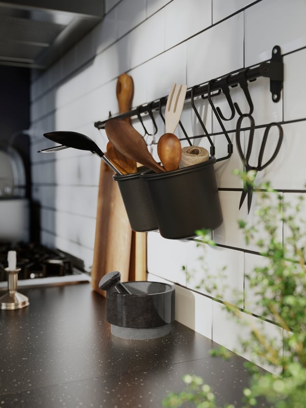 A black HULTARP rail has two containers with kitchen utensils in them and a pair of scissors hanging from one of the hooks.