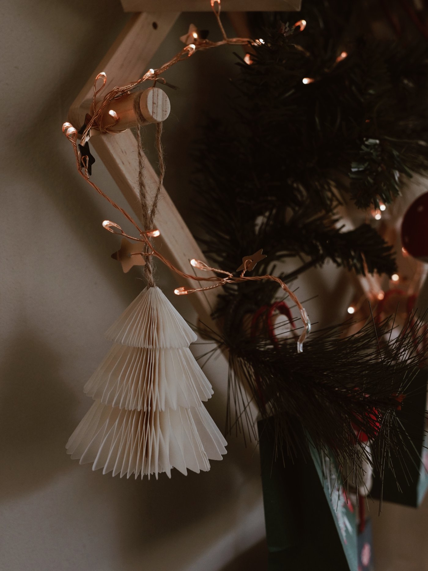 Ein gefaltetes Papier-Weihnachtsbaumornament hängt an einer Wand mit funkelnden Lichterketten. Die Szene wirkt mit gedämpfter Beleuchtung und viel Grün warm und festlich.