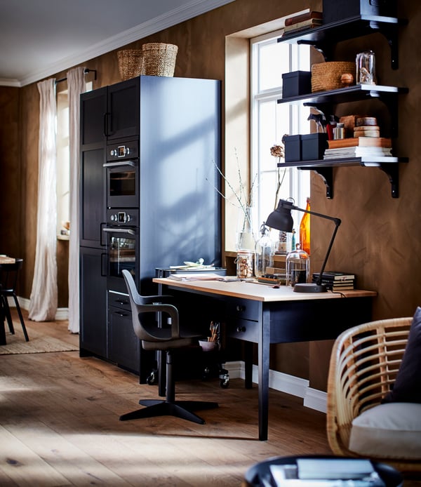 A desk in black/solid pine, a swivel chair, wall shelves in black-brown and a black kitchen section with drawers and doors.