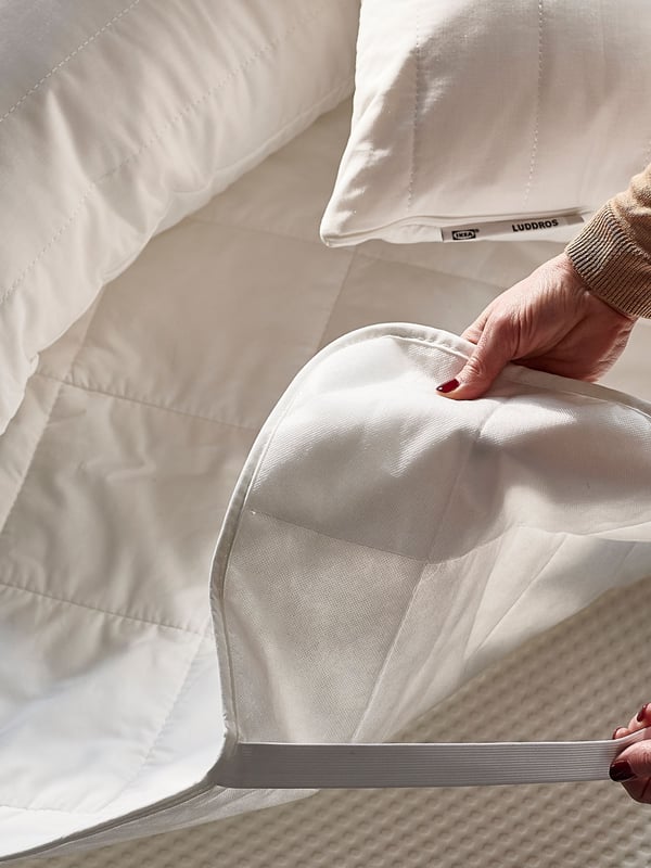 A person’s hands – one holds a corner of an ÄNGSKORN mattress protector; the other holds its elastic strap above a mattress.