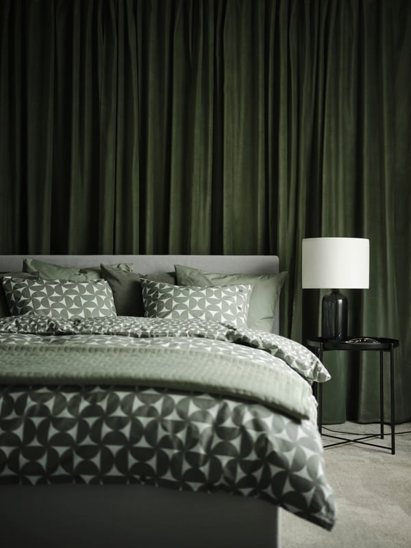 A GLADSTAD upholstered bed covered in grey/green ÄNGSNEJLIKA bed linen with a side table holding a TONVIS table lamp by it.