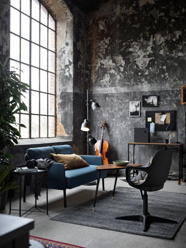 A medium-blue GLOSTAD two-seat sofa and a LÖVBACKEN side table stand beneath a big, tall window in a room with rough walls.