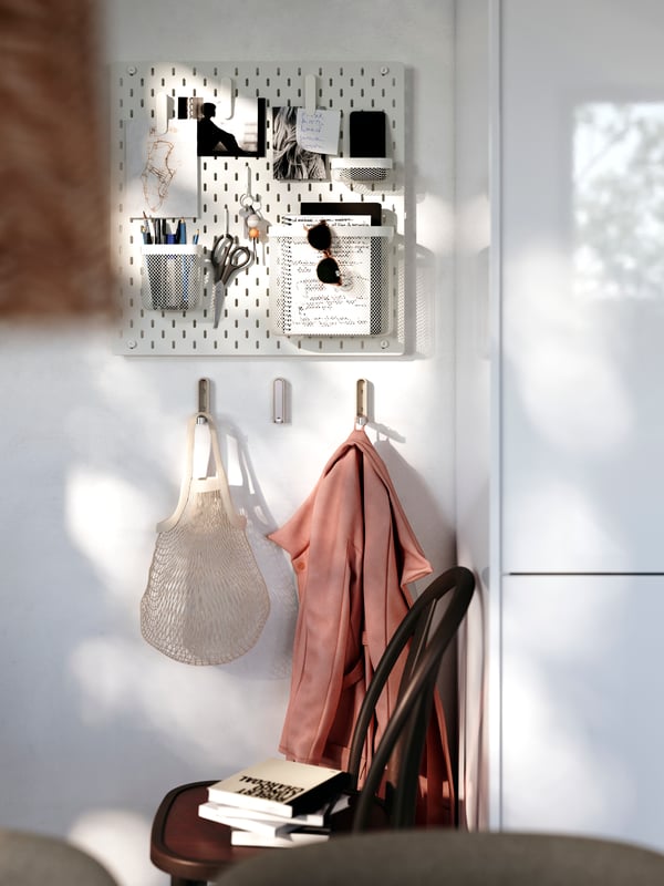 A SKÅDIS pegboard with notes and other things attached to it is placed on a kitchen wall with hooks with bags below it.