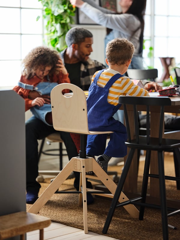 Four family members in a dining area. One child sits on a GRÅVAL birch highchair standing next to a black AGAM junior chair.