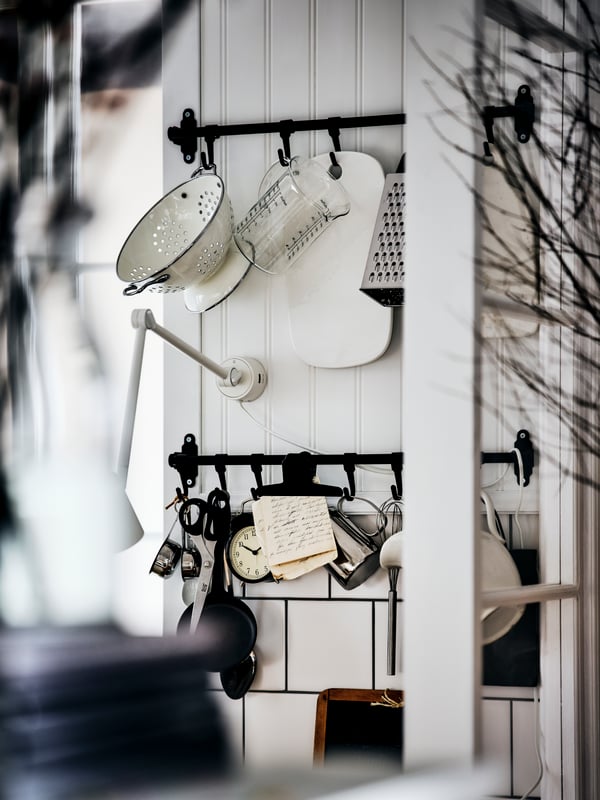 Two black HULTARP rails on a white panelled wall, with utensils like a colander, grater and jugs hanging from the hooks.