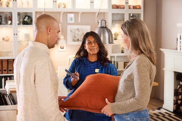 An IKEA interior designer helps two customers in a showroom, showing planning ideas on their smartphone.