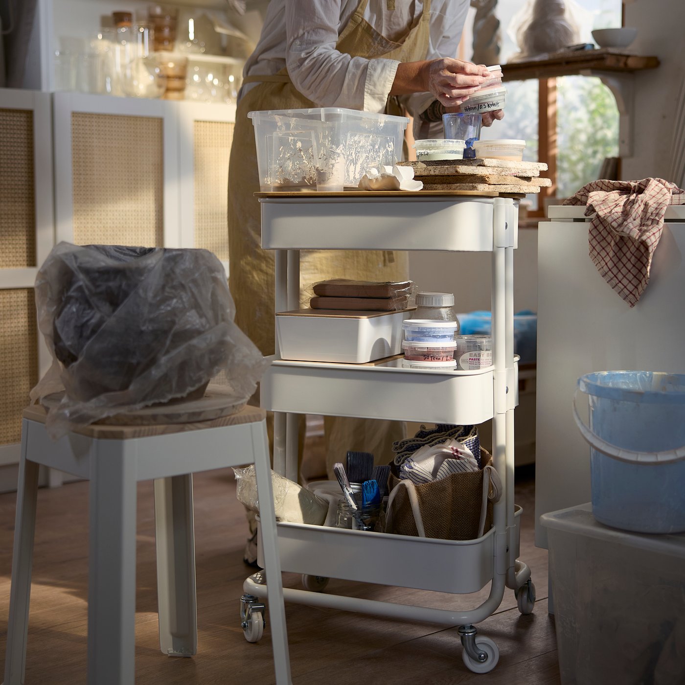A hobby space with a white RÅSKOG trolley holding tools and containers beside a person sculpting pottery in the kitchen.