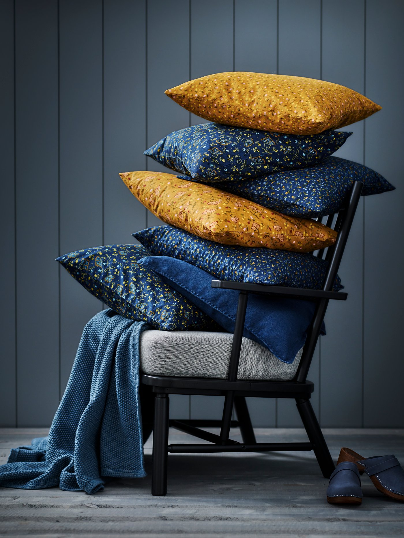 A lot of cushions in blue SISSIL cushion covers lie on top of two footstools against a wall in a run-down building.