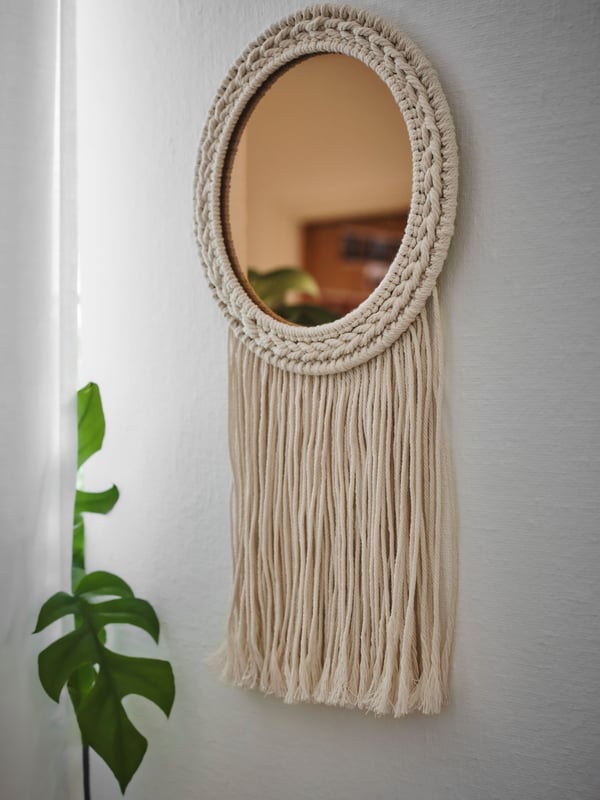 Miroir décoratif ENERGISKOG avec franges/beige couleur cuivre sur un mur à côté d’une grande plante verte et d’un rideau.