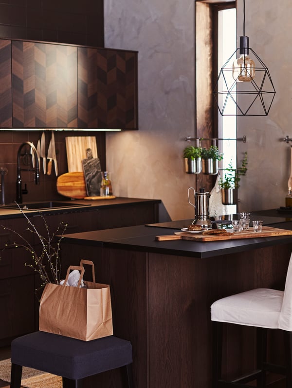 A dark wood kitchen with an island that has a BRUNSTA pendant lamp overhead. A coffee pot and glasses are on the counter.