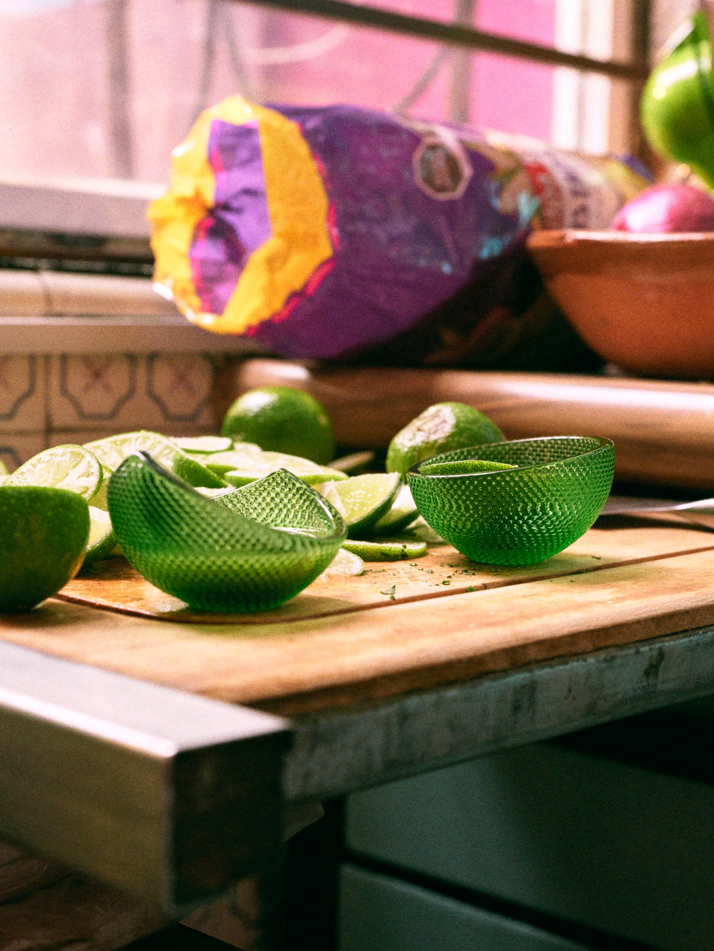 Two ÖMSESIDIG serving bowls in green glass with a relief pattern, limes and lime slices on a wooden worktop.