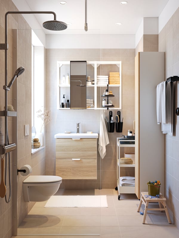 A sun-drenched bathroom in modern style with beige tiles and floors, ENHET bathroom furniture and a wall-mounted shower.