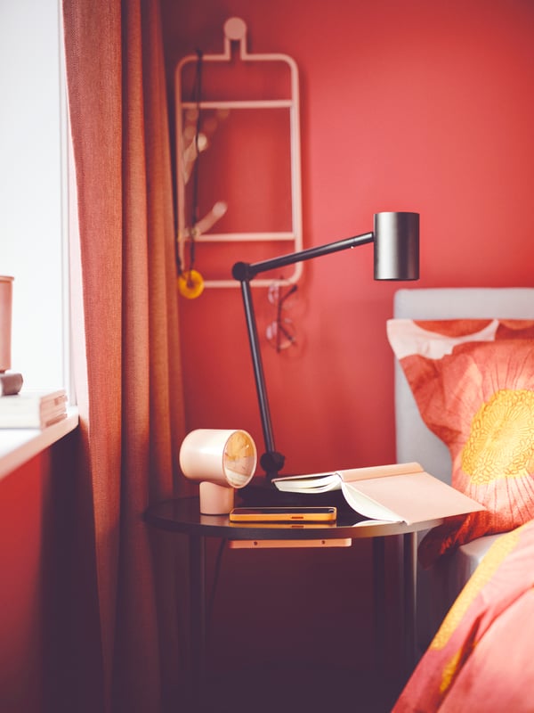 A pink BONDTOLVAN clock on an ÄSPERÖD side table, near red curtains and floral SVARTKLINT bedding and GLADSTAD bed.