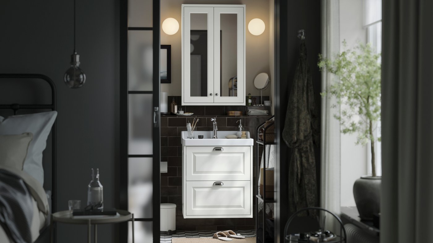 Es ist ein dunkles Zimmer das Modern eingerichtet ist. Im Hintergund kann man weiße Kästen sehen und einem Bett auf der Linken Seite mit dunkler Bettwäsche.