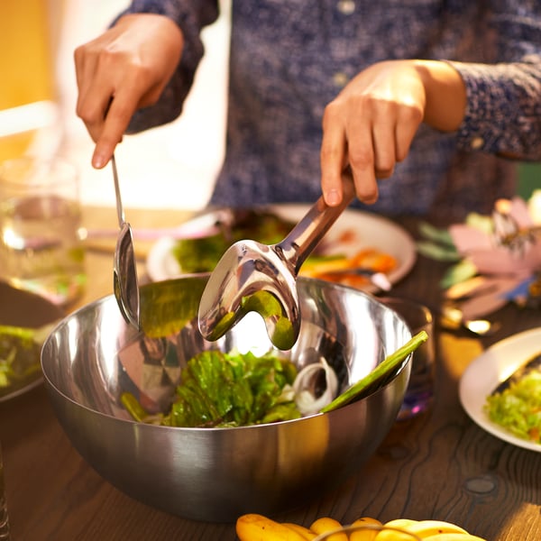 A person is tossing a salad in a large serving bowl with an ÖMSESIDIG 2-piece serving set in stainless steel.