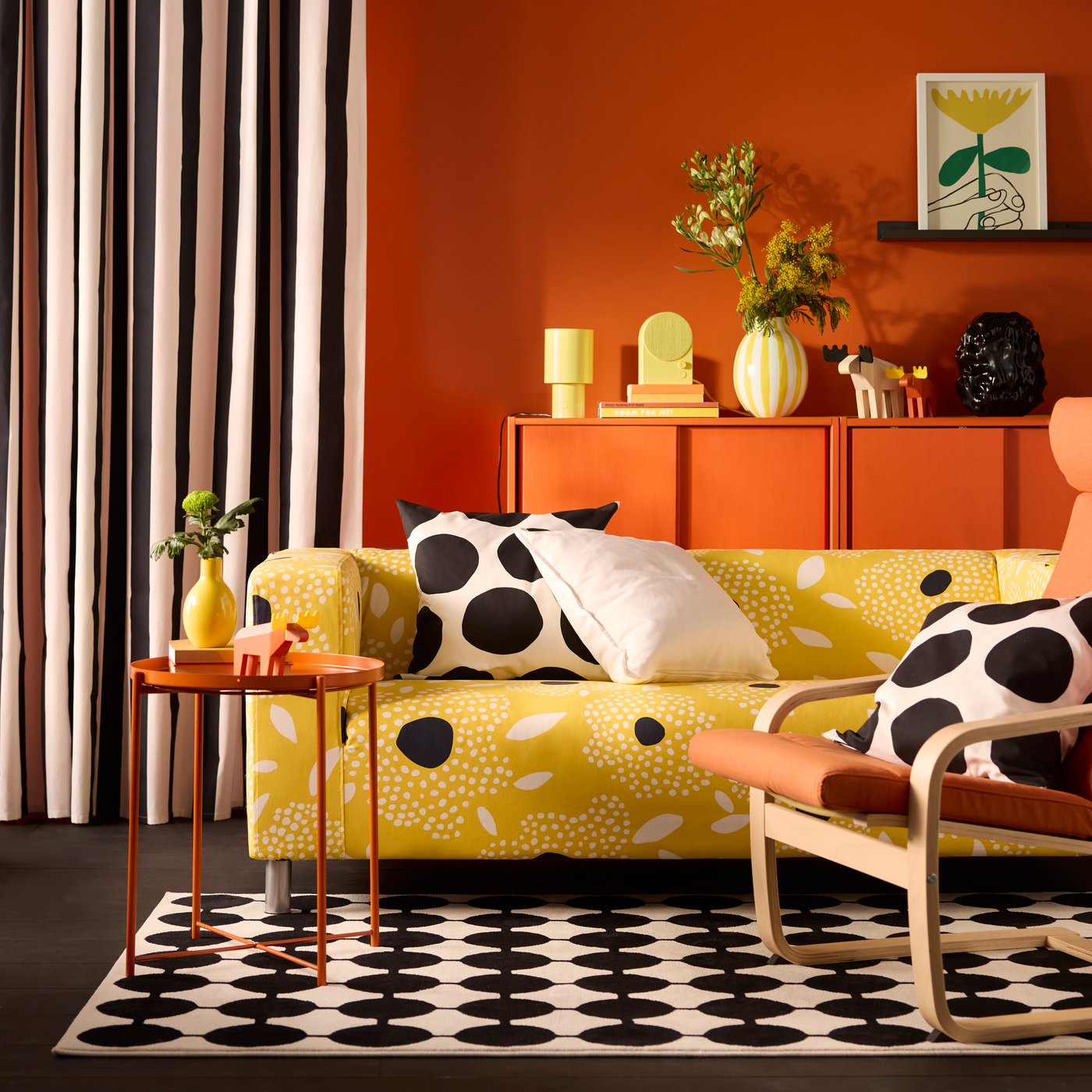 A yellow and white floral-patterned KLIPPAN cover for 2-seat sofa on a sofa in a vivid living room with an orange back wall.