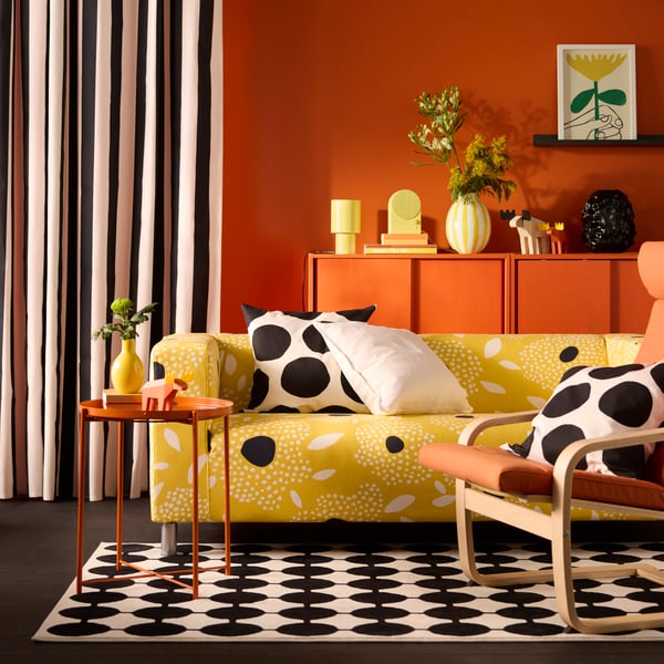A yellow and white floral-patterned KLIPPAN cover for 2-seat sofa on a sofa in a vivid living room with an orange back wall.