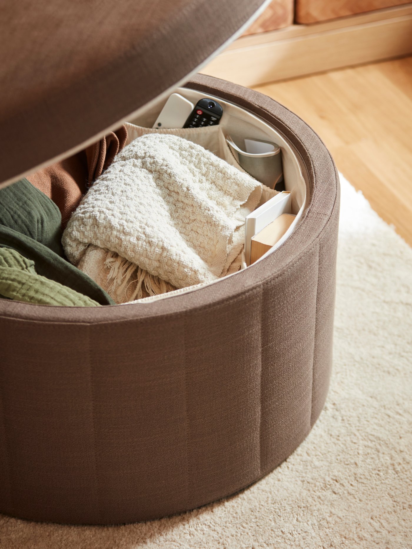 Various items, including throws and remote controls, are stored inside a SIDSJÖ pouffe with storage whose top is open.