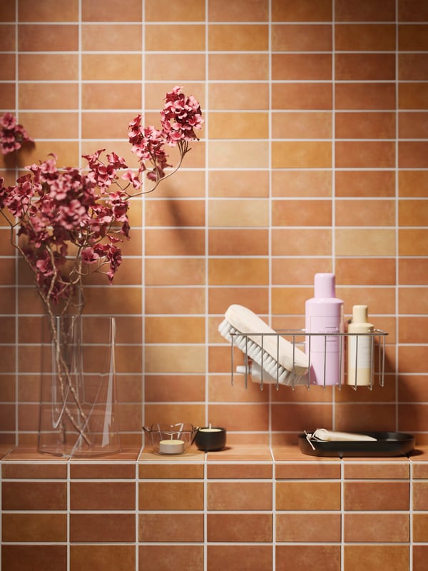 The tiled wall of a bathroom with a zinc plated KROKFJORDEN basket with suction cup with bathroom articles by a glass vase.