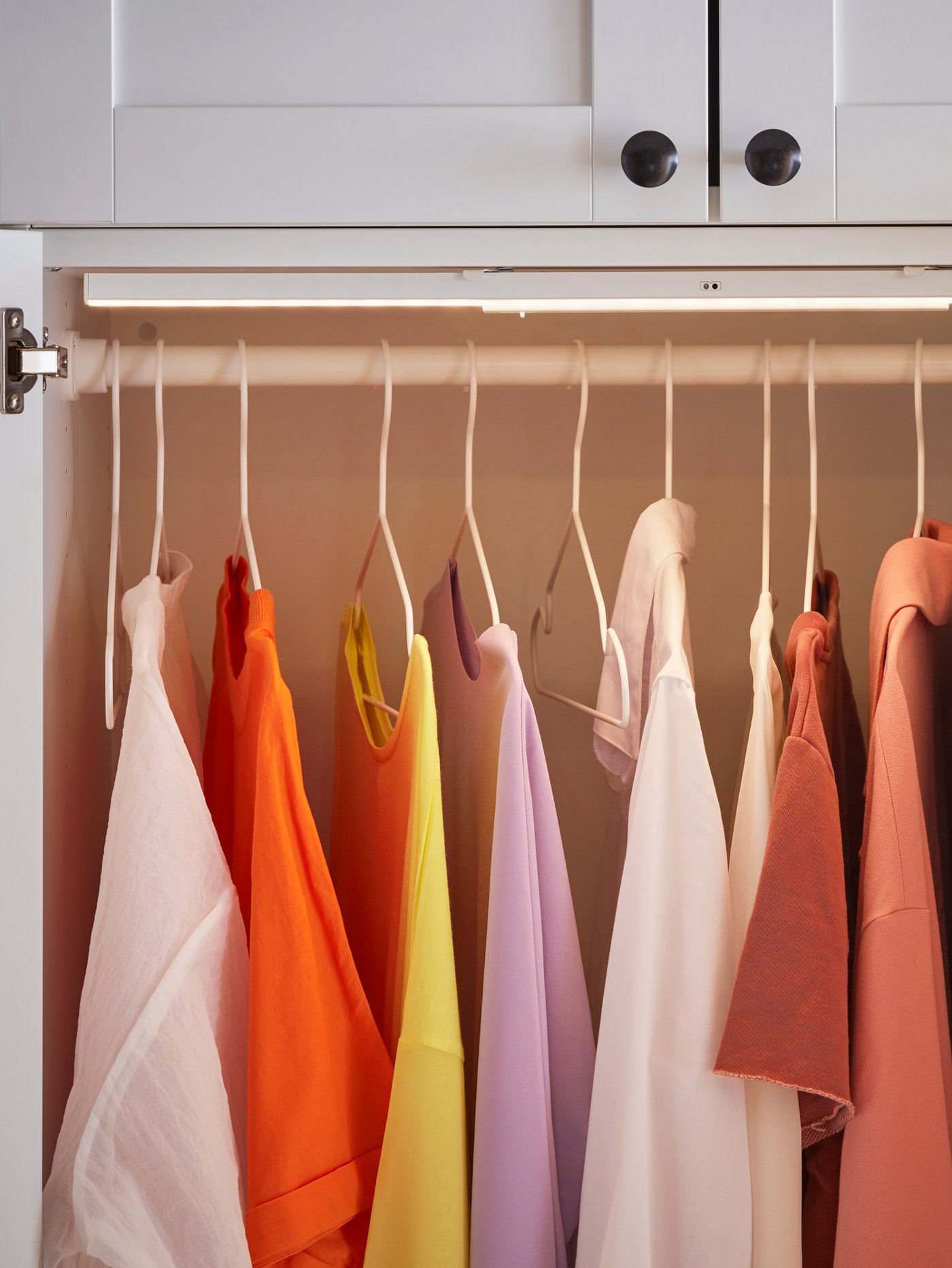 Clothes in warm tones hanging neatly on a rail inside a light-coloured wardrobe.