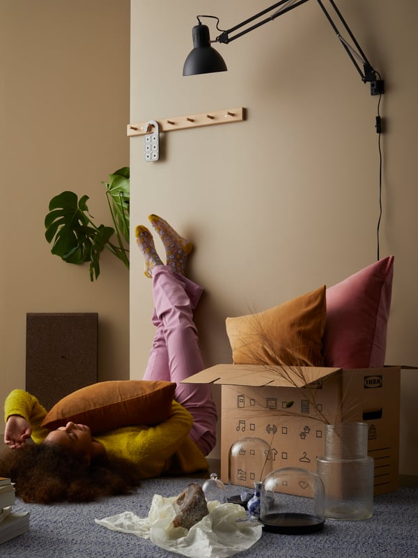 A girl lies on the floor. SANELA cushions pop out of a box, and a HÖVOLM rack and TERTIAL lamp are mounted above.