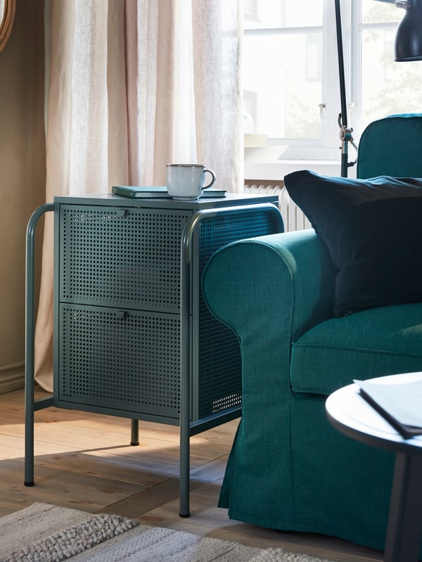 Une petite commode gris-vert à la surface perforée, un canapé turquoise foncé, un tapis rayé et des rideaux.