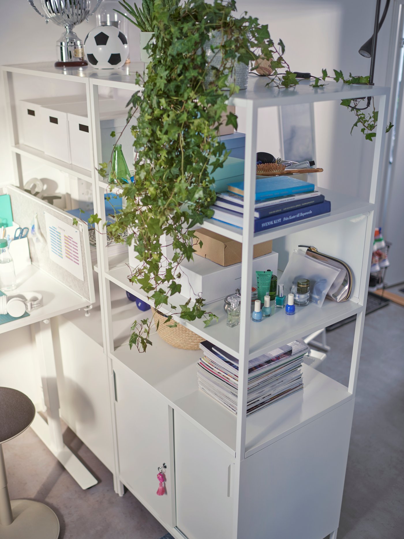 Un estante blanco con repisas abiertas en la parte superior y pueras en la parte inferior. En la parte superior hay plantas artificiales y libros.
