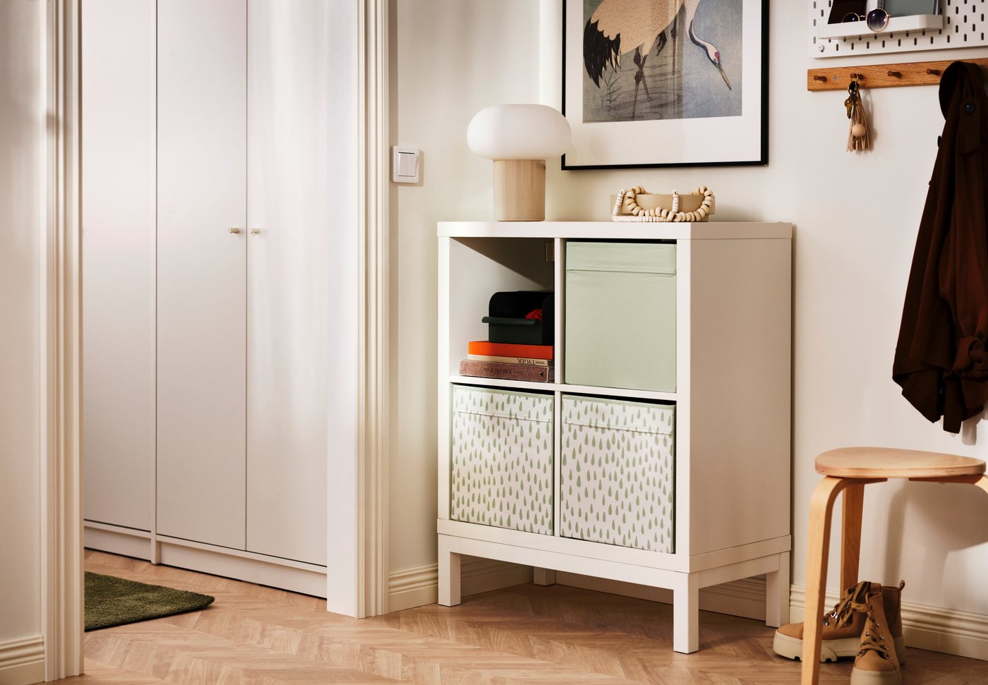 A tidy hallway featuring a white cube storage unit with patterned and solid-colored boxes. A lamp sits on top, and a wooden stool with a coat and boots is placed next to it on the herringbone floor.