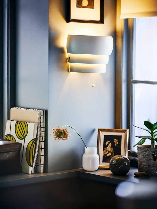 Eine eingeschaltete GRÖNPLYM Wandleuchte an einer Wand mit einem Regal darunter. Auf dem Regal steht eine Vase, die von einem gerahmten Foto und einer Topfpflanze eingerahmt ist.