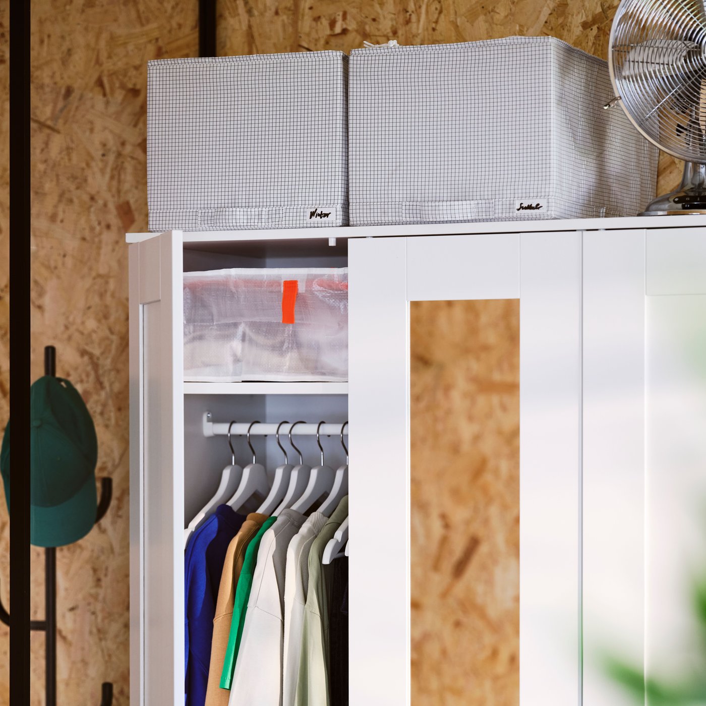 A close-up of a white BRIMNES wardrobe with storage boxes on top. On door open to show hanging shirts in neat order.