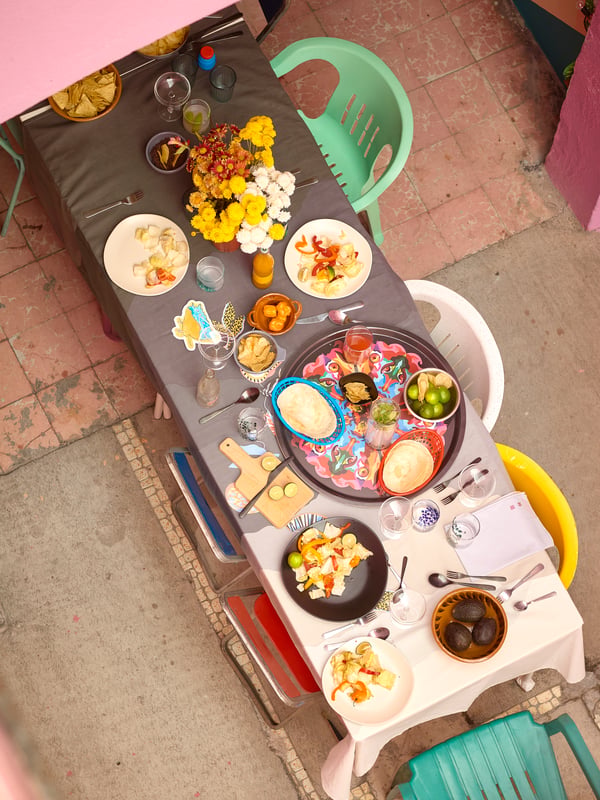 A party table with multicoloured chairs, food, flowers and a tablecloth, tray and plates from the ÖMSESIDIG collection.