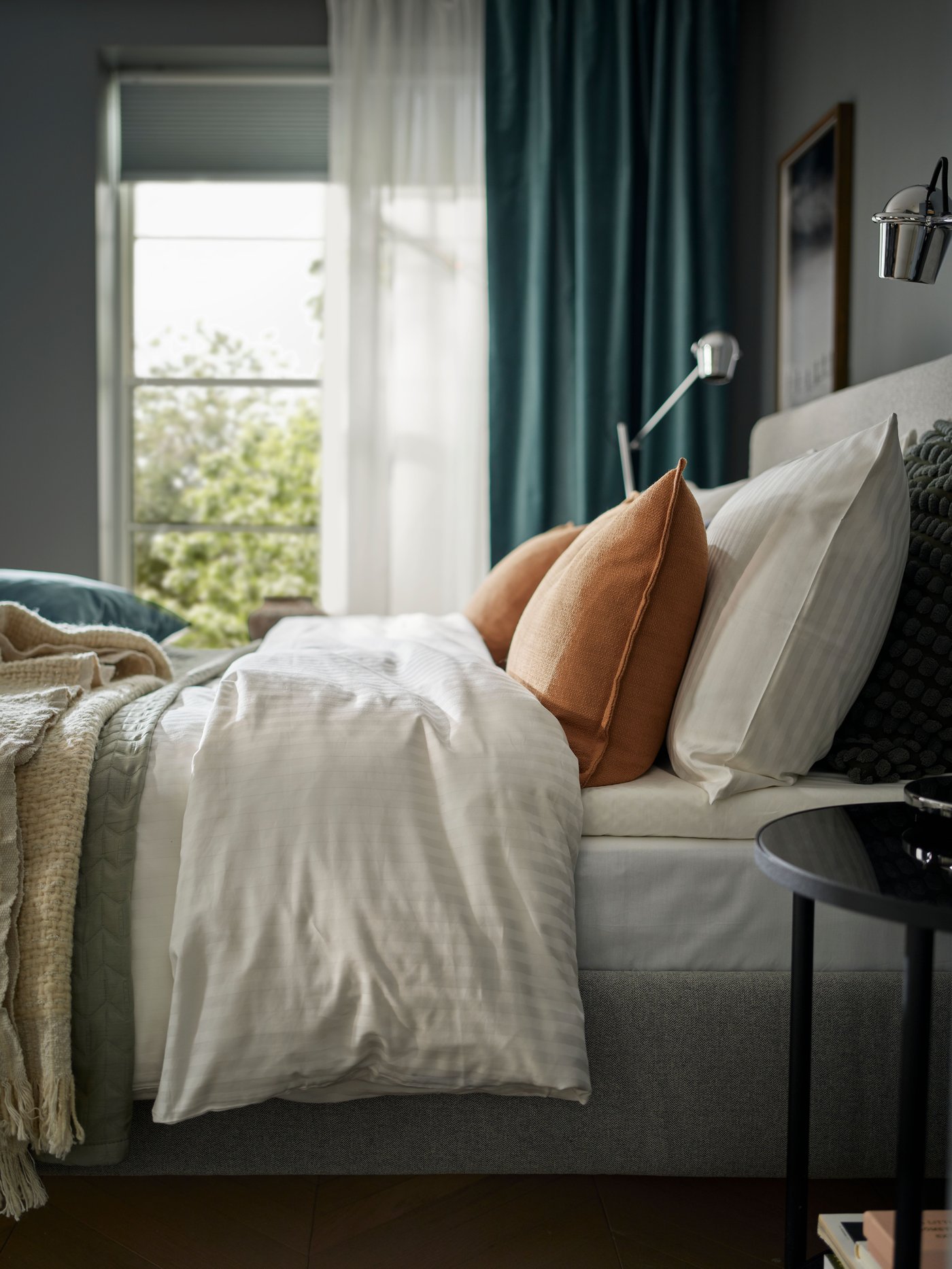 Close-up of bed shot from the side, bedding in layers of different fabrics and colours. Behind it a window with curtains.
