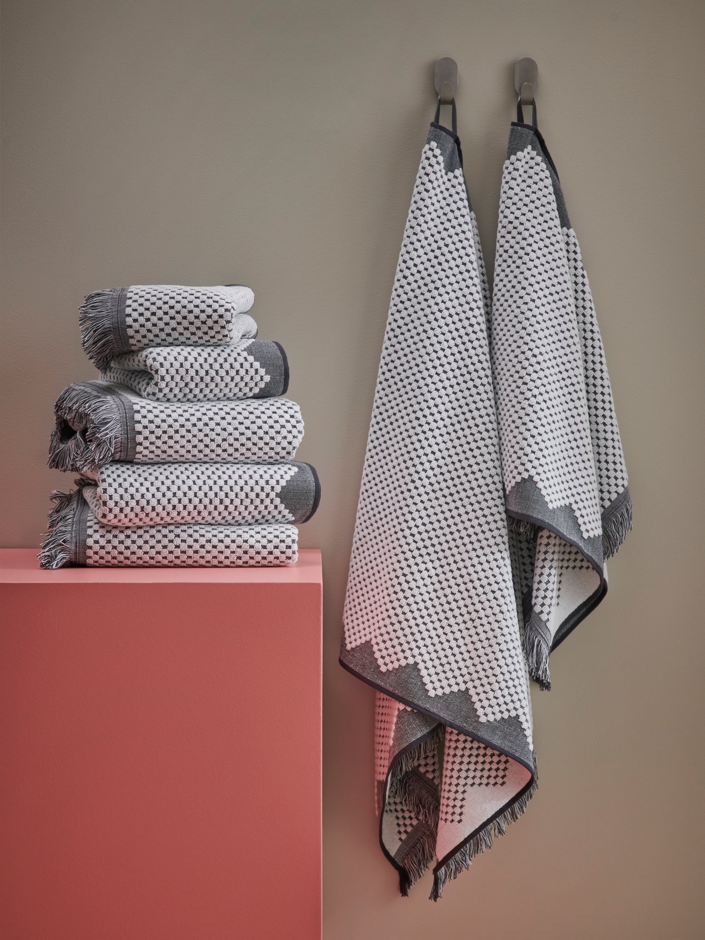 A FJÄLLSTARR bath towel and hand towel hang from hooks on a beige wall. Five towels are stacked on a pink pedestal.