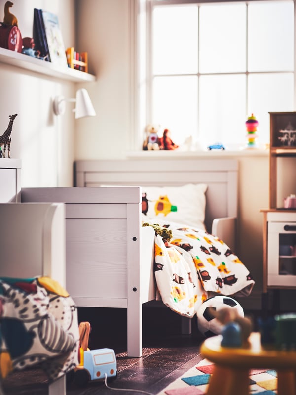 Una recámara infantil con una lámpara de pared LED FUBBLA blanca sobre una cama extensible SUNDVIK gris con un juego de edredón, almohada y ropa de cama.