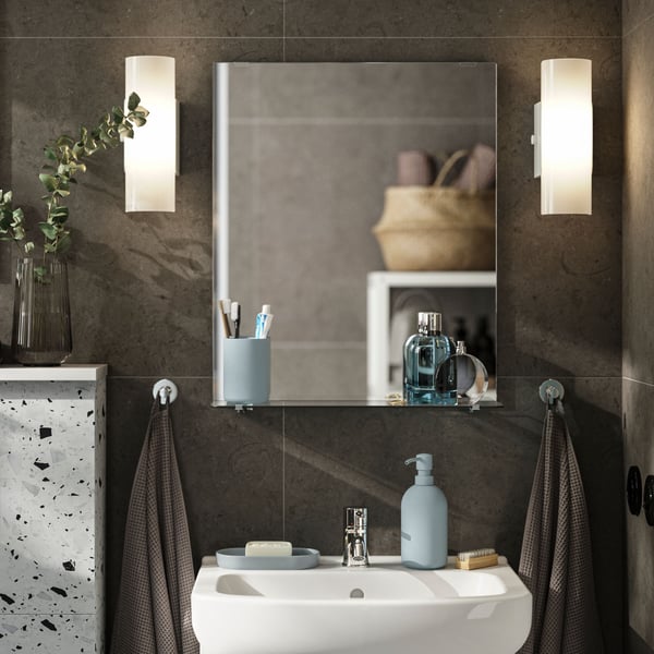 A rectangular NYSJÖN mirror with a glass shelf holds toothbrushes and perfume bottles above a white sink and cabinet.