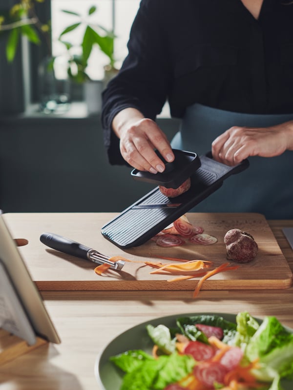 Une personne qui porte un tablier à la taille SANDVIVA qui coupe des légumes à l’aide d’une mandoline IKEA 365+ sur une planche à découper PROPPMÄTT en hêtre.