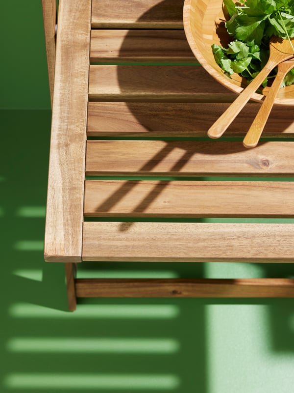 The corner of a DJUPÖN folding table, made from acacia. There is a salad and salad servers in a GRÖNSAKER serving bowl.