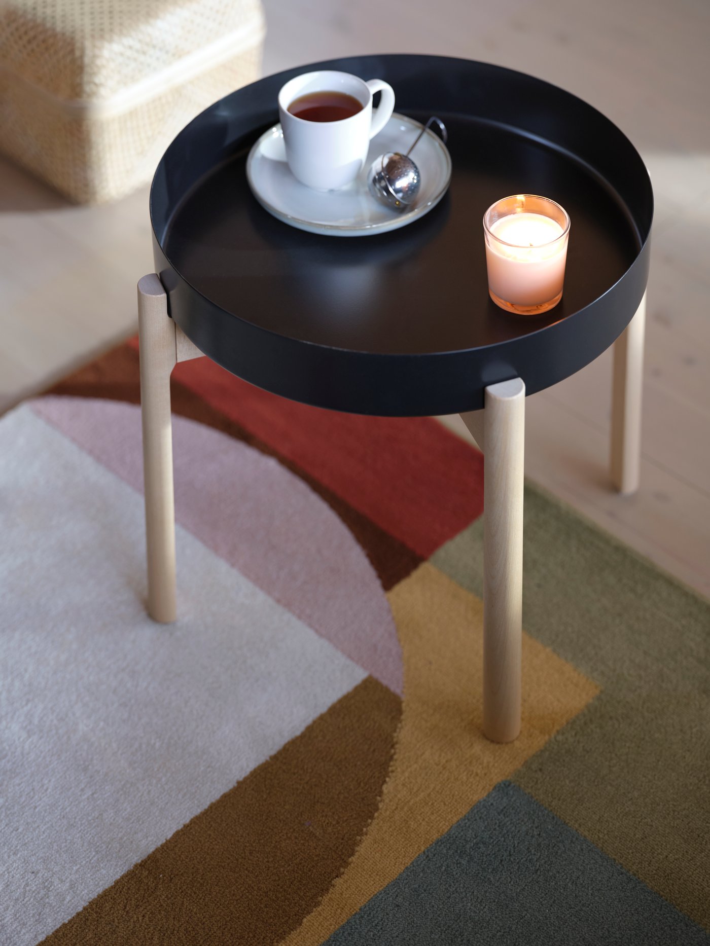 A multicoloured rug with a geometrical pattern, a black YPPERLIG coffee table with birch legs, a cup of tea and a candle.