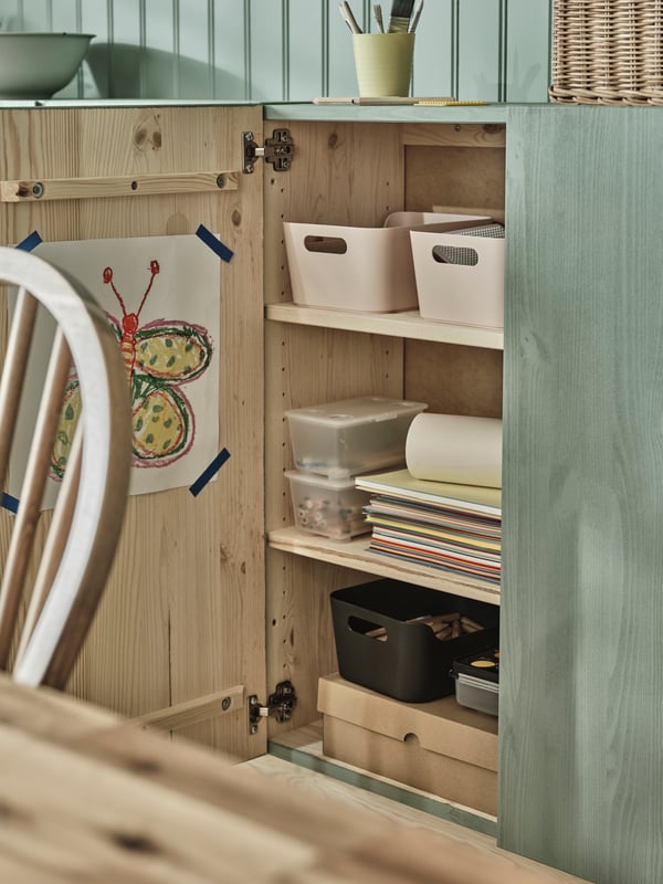 An open door to an IVAR cabinet painted in light green filled with kid’s drawings and hobby stuff, organised in boxes.