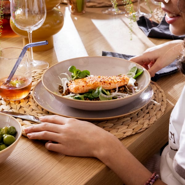 A person sitting at a TONSTAD table, about to eat food in a FÄRGKLAR bowl standing on a FÄRGKLAR plate, and STORSINT glasses.